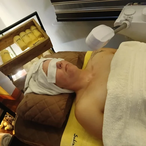 Facial treatment being performed on a client lying on a spa bed with a towel covering the body and a facial steamer positioned above.